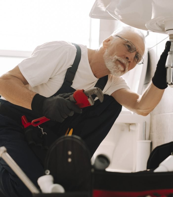 Senior plumber wearing blue overalls is on his knees, tightening the pipe under a bathroom sink using an adjustable wrench